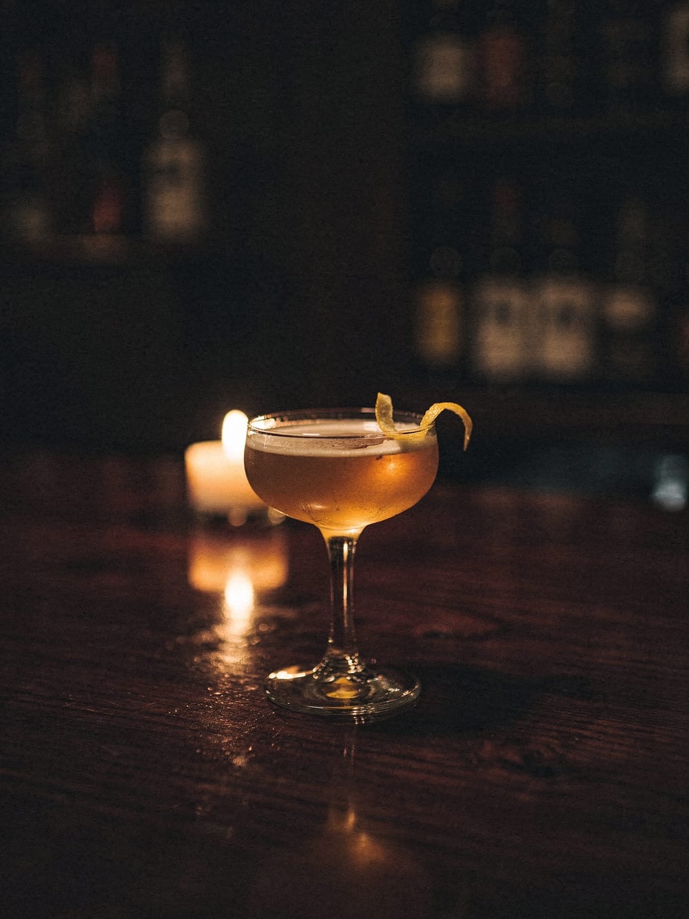 Golden cocktail in a coupe glass on a dark wood bar in candlelight